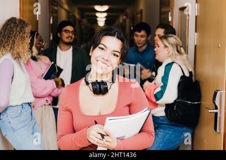 Portrait of smiling young woman with book while multiracial friends in background at corridor in college dorm Stock Photo