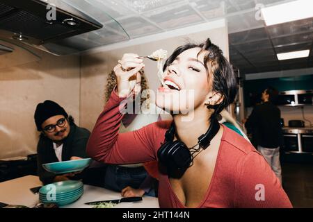 Happy young woman eating noodles by friend in kitchen at college dorm Stock Photo