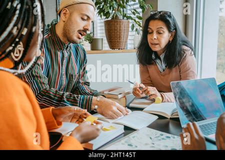 Multiracial students discussing over book while studying together at college cafeteria Stock Photo