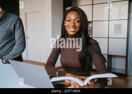 Portrait of smiling young female student sitting with book studying by friend at community college Stock Photo