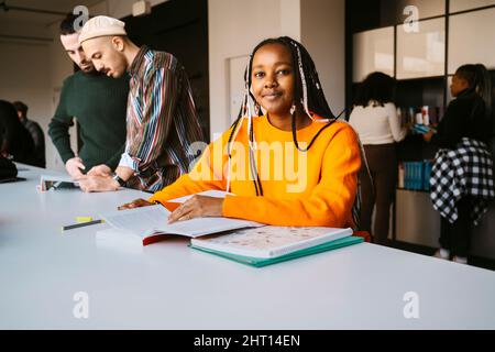 Portrait of smiling woman with braided hair studying by friends discussing in community college Stock Photo