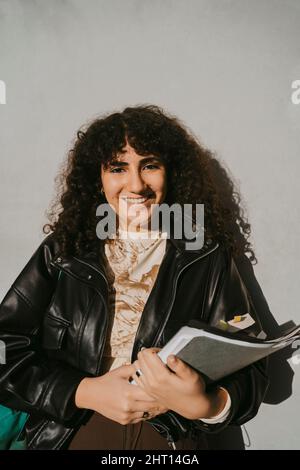 Portrait of smiling young woman wearing leather jacket holding book leaning on gray wall Stock Photo