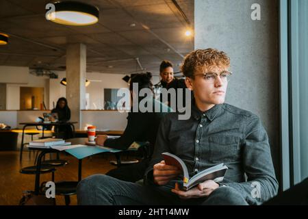 Thoughtful young blond man sitting with book in university cafeteria Stock Photo