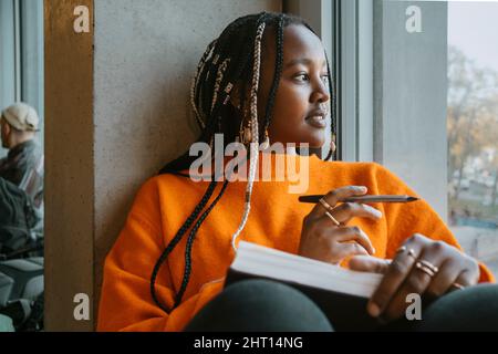 Thoughtful young woman with braided hair looking out through window in university cafeteria Stock Photo