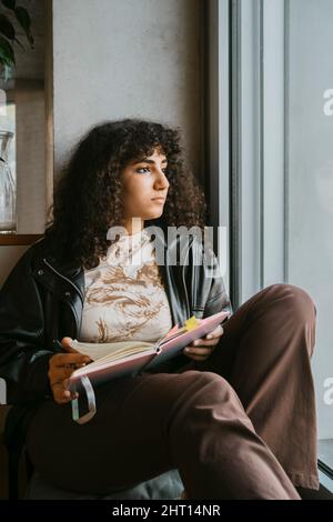Thoughtful young woman sitting with book looking out of window at university Stock Photo