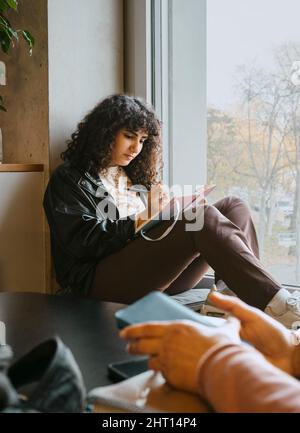 Young woman writing in book while sitting by window at university Stock Photo