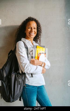 Portrait of smiling woman holding book and backpack leaning on gray wall at college Stock Photo
