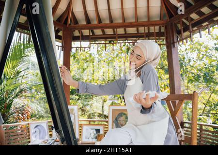 muslim asian female artist painting on canvas and talking to phone Stock Photo
