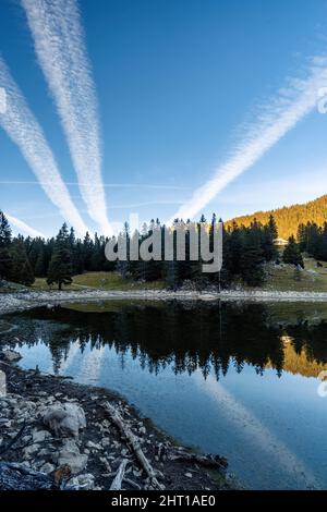 An alpine lake in the Swiss Alps in early autumn and just before sunset. The water is calm and reflects the fir forest on the other side, In the sky there are 3 trails of planes just passed by. Stock Photo