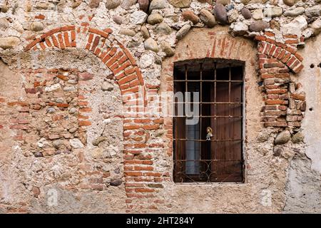 Rocky facade of a medieval house of Bassano del Grappa with a barred window, a small doll placed in the window. Stock Photo