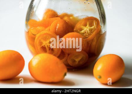 Sliced kumquat fruit in syrup water in glass, whole fruit on white background, closeup. Stock Photo