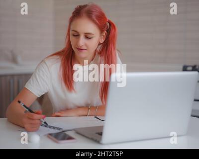 freelancer working on computer at home, making notes in notebook while listening to lecture or webinar. Attractive woman studying online using laptop software, web surfing or online shopping. Stock Photo