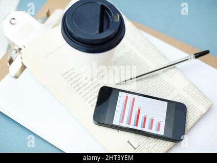 Keeping an eye on the news and the stock market. Smartphone with bar graph on screen sitting ontop of a newspaper and clipboard with a disposable cup Stock Photo