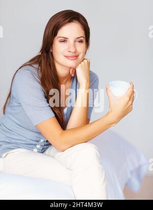 Thoughtful female drinking a cup of coffee at the bedroom. Day dreaming - Pretty casual female drinking coffee and sitting relaxed on the bed. Stock Photo