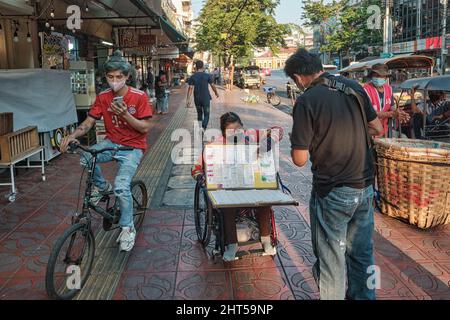 On a sidewalk in Bangkok, Thailand, a handicapped lottery vendor in a wheelchair serves a customer, while a cyclist engrossed in his mobile rides by Stock Photo