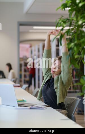 Tired woman adult learner stretching arms with closed eyes while studying in cozy library Stock Photo