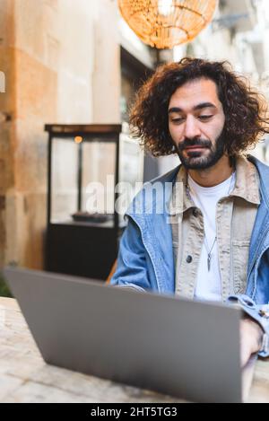 Bearded Hispanic male freelancer in casual clothes with curly hair sitting at table and browsing data on netbook while working in street cafeteria Stock Photo
