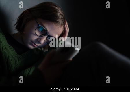 Sad little girl, alone in darkness, sitting and using smartphone. Stock Photo