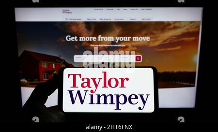 Person holding smartphone with logo of British housebuilding company Taylor Wimpey plc on screen in front of website. Focus on phone display. Stock Photo