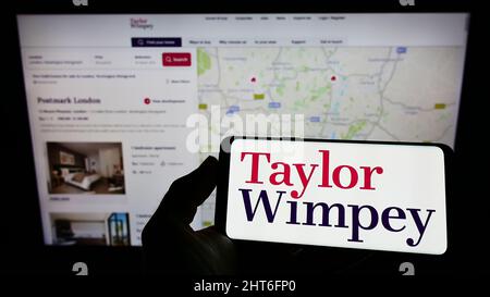 Person holding mobile phone with logo of British housebuilding company Taylor Wimpey plc on screen in front of web page. Focus on phone display. Stock Photo