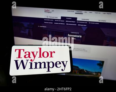 Person holding cellphone with logo of British housebuilding company Taylor Wimpey plc on screen in front of webpage. Focus on phone display. Stock Photo