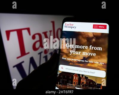 Person holding cellphone with website of British housebuilding company Taylor Wimpey plc on screen with logo. Focus on center of phone display. Stock Photo