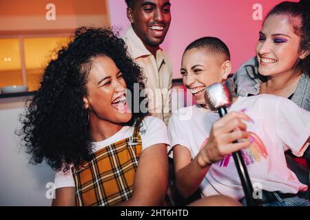 Ecstatic friends enjoying karaoke night. Happy young friends laughing joyfully while singing their favourite song at a house party. Group of multicult Stock Photo