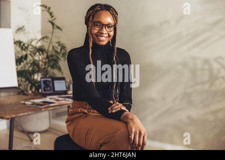 Cheerful female freelancer looking at the camera while working on a new project in her home office. Happy young photographer sitting on a chair with h Stock Photo