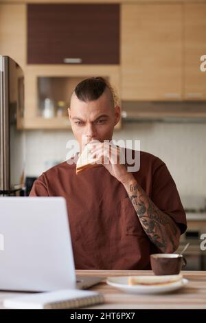 Young man eating sandwich and drinking coffee when reviewing programming code on laptop screen Stock Photo