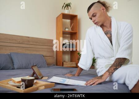 Pensive young marketing manager in bathrobe sitting on bed and analyzing data in reports Stock Photo