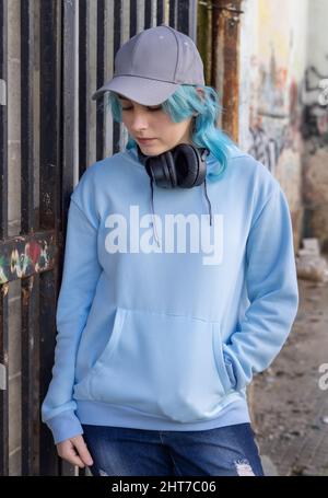 Teenager wearing light blue oversize hoodie and baseball cap looking down. Blue haired teen girl with wireless headphones stays outdoor near a gate. C Stock Photo