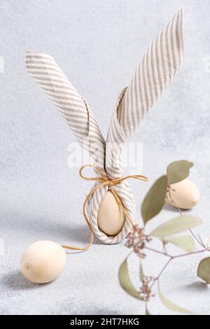 Easter eggs wrapped in linen napkin in form of bunny with ears, olive branch. Easter decor in pastel colors on gray textured background. Traditional Stock Photo