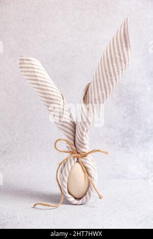 Egg wrapped in linen striped napkin tied with jute in form Easter bunny with ears form on light background. Minimalism concept, vertical Easter card Stock Photo