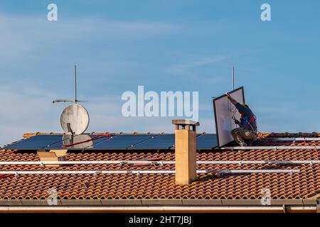 A technician lifts a solar panel as he is installing a photovoltaic system on top of a red-tiled roof Stock Photo
