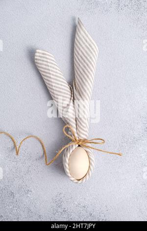 Wooden Egg wrapped in linen striped napkin tied with jute in form Easter bunny with ears form on light gray background. Minimalism concept, vertical Stock Photo