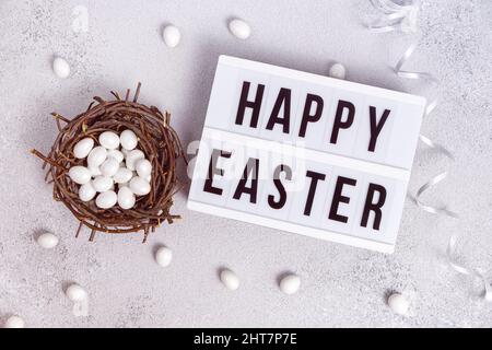 Traditional wish Happy Easter on a light board. Candies in form of chocolate eggs in white icing in bird's nest. Egg hunting. Light gray gradient Stock Photo