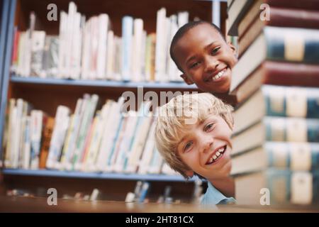 Hello. Two school friends peering around a stack of books in the library. Stock Photo
