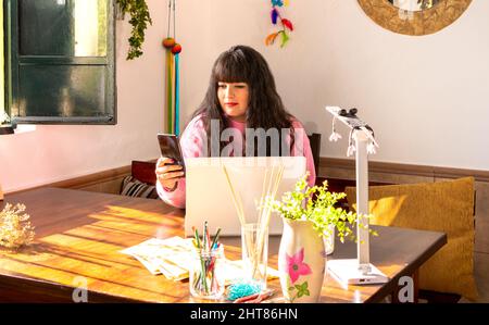 Brunette young woman using smartphone and laptop in sunny home office. Stock Photo