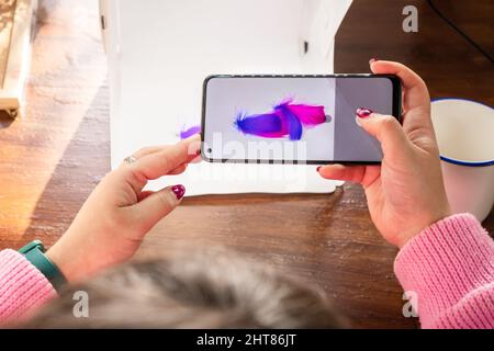 Freelancer anonymous female artisan taking picture of handmade feather earrings for social media blog while using smartphone and sitting at table in w Stock Photo