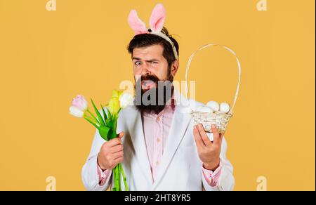 Prepare for Easter. Bearded man in bunny ears with bouquet of tulips and basket eggs. Discount. Sale. Stock Photo