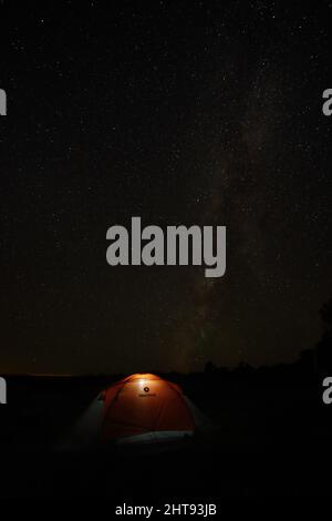 View of the Milky Way and night sky with a brightly lit orange tent at a camp on the South Rim of the Grand Canyon. In the distance the lights of Came Stock Photo