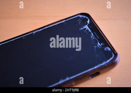 Black smartphone with cracked screen protector, isolated on a brown wood background. Landscape orientation. Stock Photo