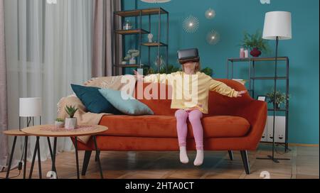 Toddler girl sitting on home sofa using virtual reality headset helmet app to play simulation game Stock Photo