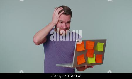 Exhausted man freelancer with pasted sticker notes using laptop computer having concentration problem creative crisis. Nervous adult guy confused by b Stock Photo
