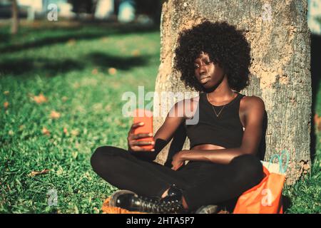 Portrait of a woman sitting cross-legged on the grass, leaning against the tree and holding a cellphone with one hand, a sunny day. Black curly-haired Stock Photo