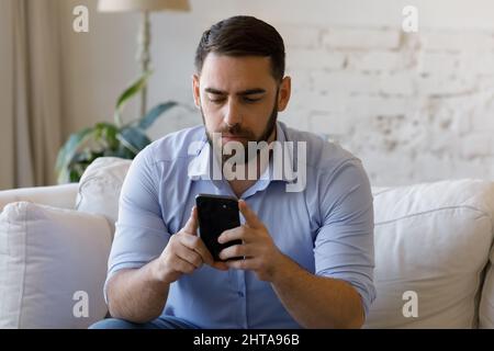 Serious man sit on couch look at cellphone screen Stock Photo