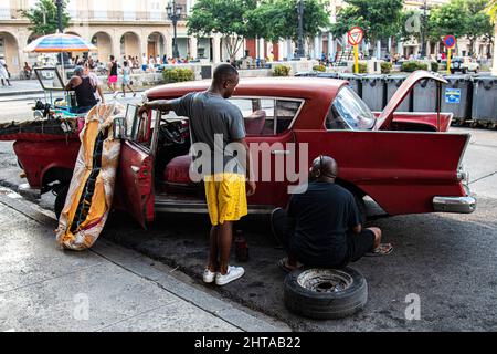 Two Cuban men talk while working on fixing a tire on a car in Havana, Cuba with a mattress on the hood of an old American classic car. Stock Photo