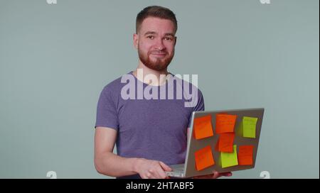 Exhausted man freelancer with pasted sticker notes using laptop computer having concentration problem creative crisis. Nervous adult guy confused by b Stock Photo