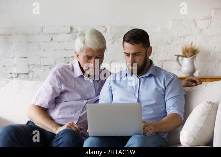 Serious millennial man helps to older father with laptop usage Stock Photo