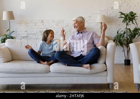 Mature grandfather and little grandson laughing seated cross-legged on sofa Stock Photo